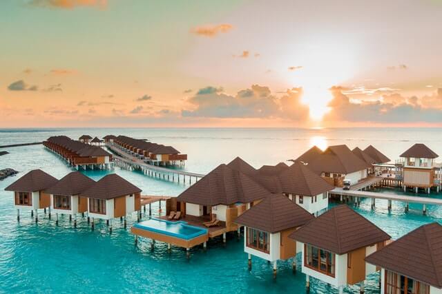 A collection of small white huts with brown roofs suspended above clear water at sunset in the Maldives
