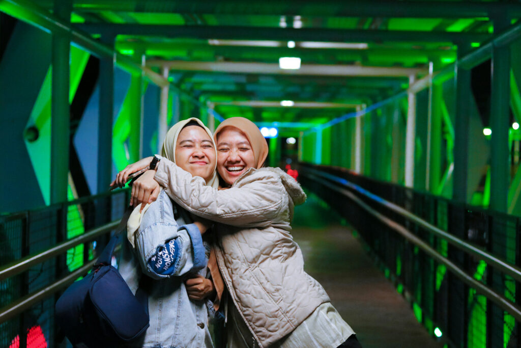 Two young women both wearing hijabs, hug each other on a green-lit bridge while laughing and smiling happily
